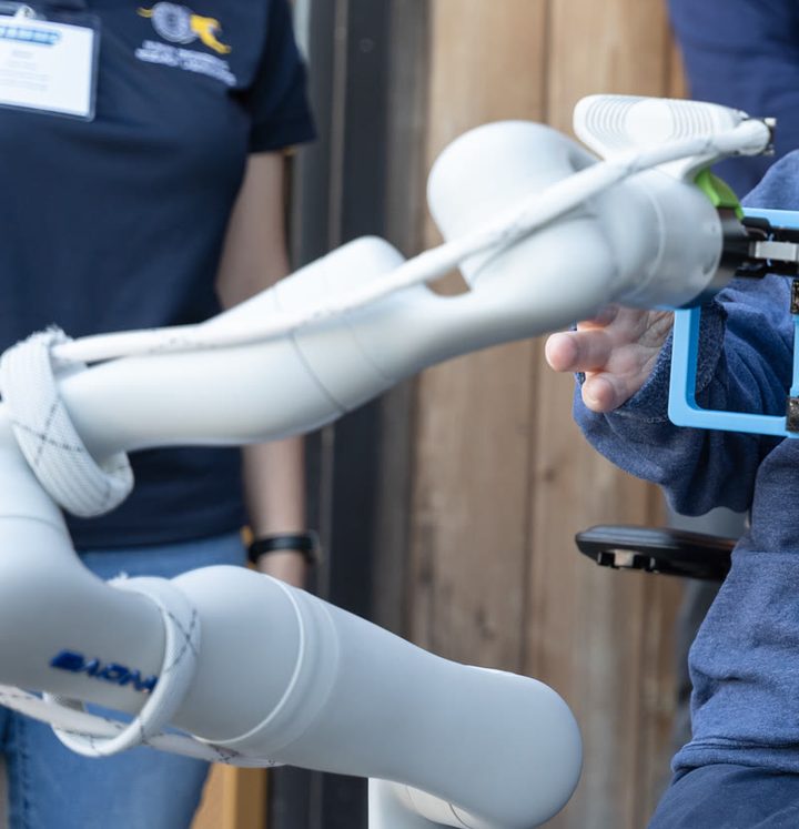 A white robotic arm holding a cup out toward a wheelchair user’s hand.