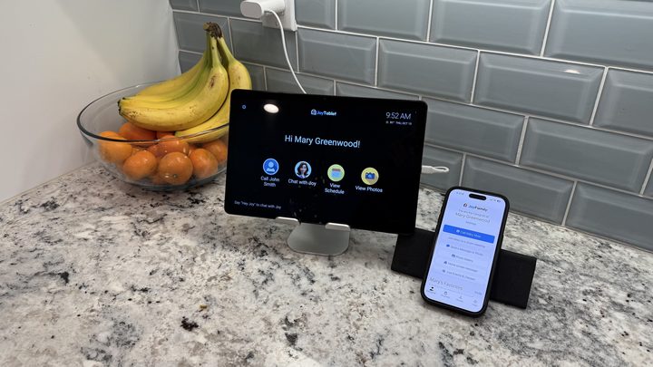 A tablet on a stand showing the JoyTablet home screen with a greeting and four large buttons for calling, chatting with Joy, viewing the schedule and viewing photos, beside a phone showing the JoyFamily app, on a kitchen counter.