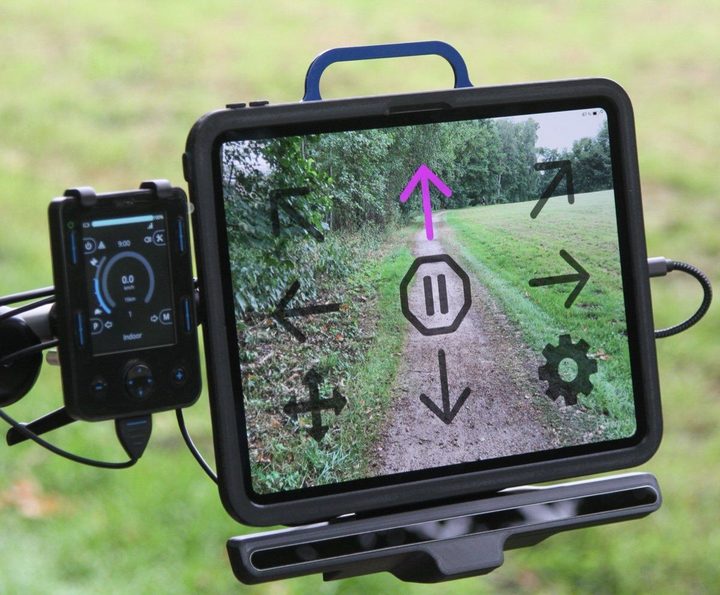 Tolt Technologies A13i: an iPad with an eye-tracker bar beneath it, showing Ability Drive direction arrows, mounted beside a power wheelchair joystick display.