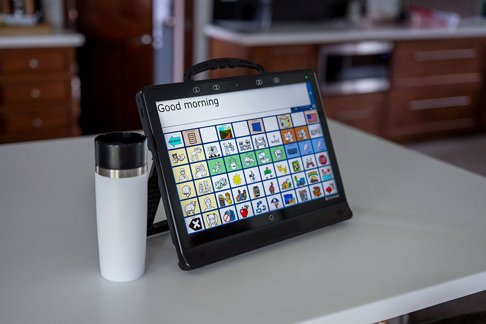 PRC-Saltillo Genesis AAC speech-generating device: a black rugged tablet on a kickstand on a countertop, touchscreen showing a vocabulary grid with Good morning in the message window; no people in the photo.